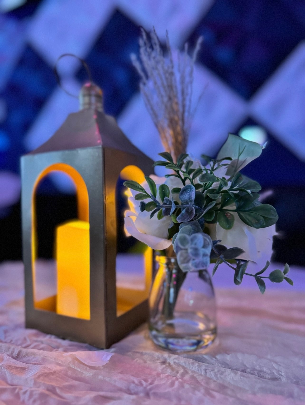 Gold lantern and white roses on a blush table runner under blue uplighting